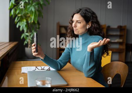 Sorridente giovane donna d'affari in auricolari seduti alla scrivania dell'ufficio parlando con videochiamata sullo smartphone Foto Stock