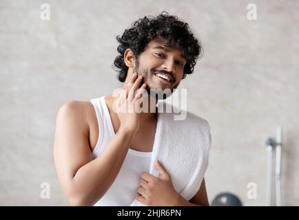 Ritratto di uomo indiano felice toccando la barba e sorridendo alla macchina fotografica, ragazzo giovane attraente godendo la routine del mattino Foto Stock