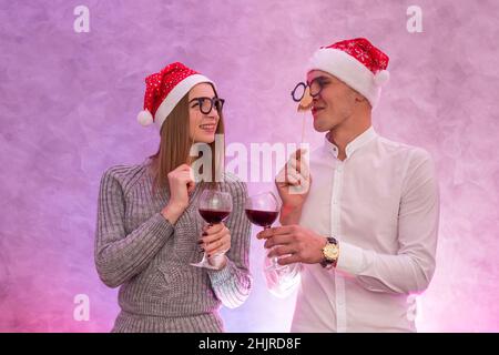 Felice coppia a babbo natale cappelli con vino in bicchieri che tengono maschera vicino al viso e festeggia San Valentino Foto Stock