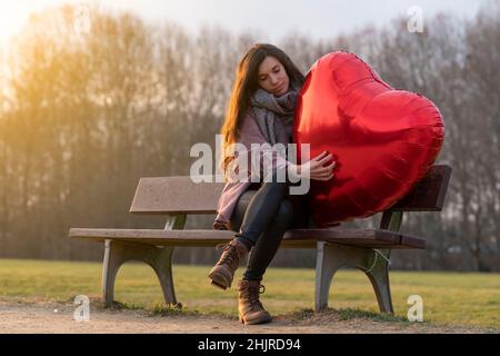 Triste giovane donna che abbraccia un pallone a forma di cuore seduto su una panchina nel parco Foto Stock