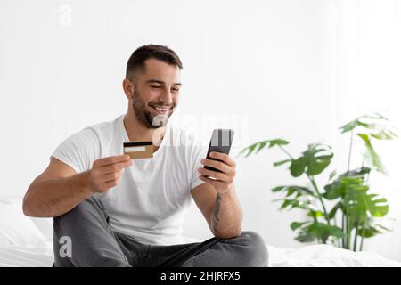 Sorridente giovane ragazzo europeo in t-shirt bianca shopping online, ordina merci su smartphone e mostra carta di credito Foto Stock