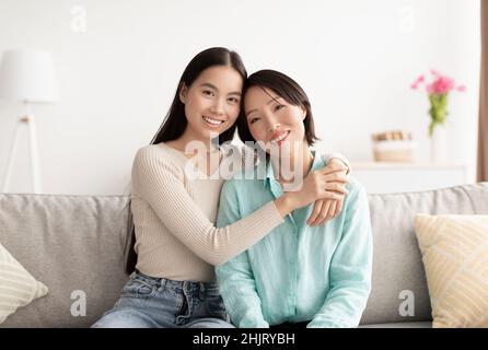 Ritratto della giovane donna asiatica e della madre matura che abbraccia e sorride alla macchina fotografica, seduto sul divano a casa Foto Stock