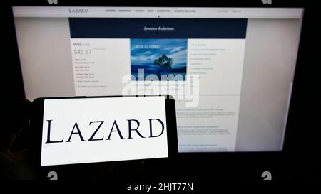 Persona che tiene il telefono cellulare con il logo della società americana di servizi finanziari Lazard Ltd. Sullo schermo di fronte alla pagina web. Mettere a fuoco sul display del telefono. Foto Stock