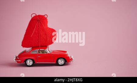 Auto rossa con un grande cuore morbido in forma di regalo sul tetto su sfondo rosa, copia space.Concept di un viaggio romantico di San Valentino o il matrimonio Foto Stock