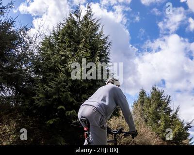 Vista ad angolo basso di un uomo anziano in pantaloni grigi e una felpa in bicicletta su un sentiero in una giornata soleggiata e nuvolosa nel Pacifico nord-occidentale Foto Stock