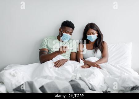 L'uomo e la donna afro-americani infelici millenni in maschere protettive guardare termometro, soffrono di ammalarsi a letto Foto Stock