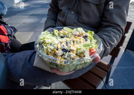 Maschio caucasico in una giacca invernale, tenendo un'insalata sud-occidentale in un contenitore di plastica da andare a un tavolo all'aperto. Foto Stock