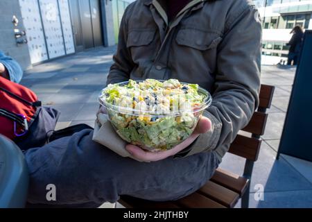 Maschio caucasico in una giacca invernale, tenendo un'insalata sud-occidentale in un contenitore di plastica da andare a un tavolo all'aperto. Foto Stock