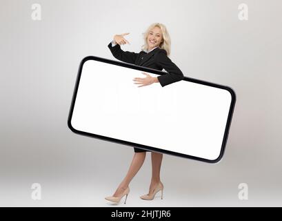 Scatto a tutta la lunghezza del corpo di una donna d'affari felice che trasporta un grande smartphone con schermo vuoto e punta allo schermo del gadget Foto Stock