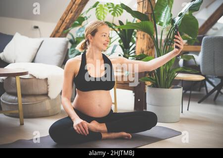 Giovane felice e allegra bella donna incinta prendendo selfie con il suo cellulare, pur rimanendo in forma, sportiva e attiva sul suo congedo di maternità Foto Stock
