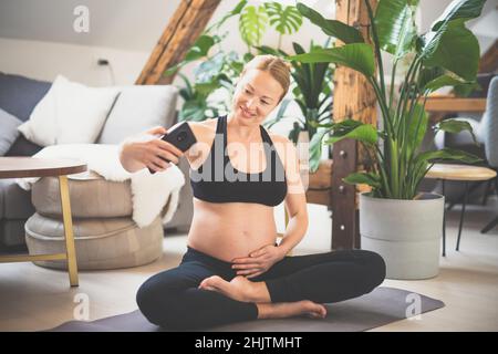 Giovane felice e allegra bella donna incinta prendendo selfie con il suo cellulare, pur rimanendo in forma, sportiva e attiva sul suo congedo di maternità Foto Stock