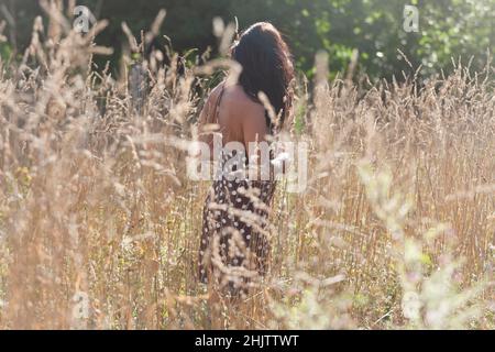 Una ragazza dalla parte posteriore con capelli lunghi e un abito marrone con pallini bianchi in un campo di erba secca alta in estate Foto Stock