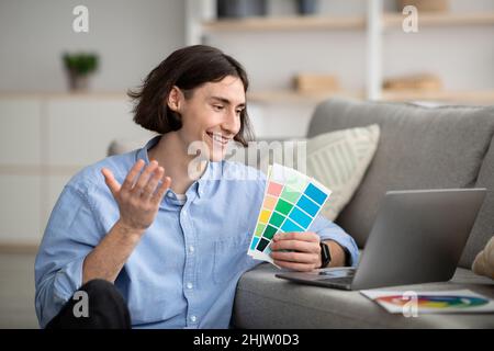 Giovane freelance maschile che tiene i campioni di colore di fronte al computer portatile, presentando il progetto grafico di design Foto Stock