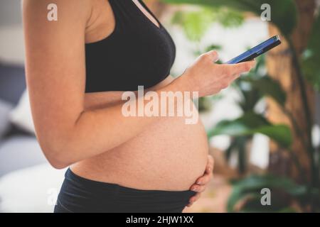 Gravidanza e tecnoloy supportato sano stile di vita concetto. Allegra donna incinta felice utilizzando l'applicazione per smartphone a casa. Foto Stock