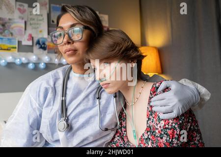 Giovane infermiera in abbigliamento protettivo che abbraccia la triste ragazza malata, lei la sostiene durante la malattia al reparto ospedaliero Foto Stock