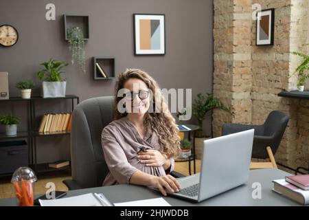 Giovane donna d'affari di successo che tiene la mano sul ventre incinta mentre si siede dal posto di lavoro e che vi guarda con il sorriso Foto Stock