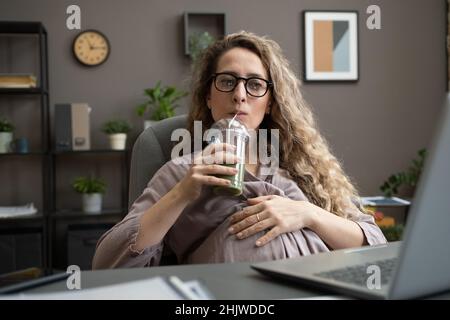 Donna d'affari incinta che beve un frullato mentre si siede dalla scrivania davanti al laptop e si guarda il video online Foto Stock