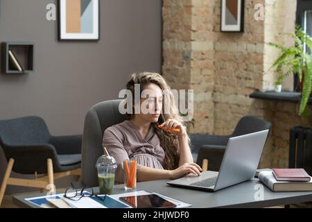 Giovane donna gravida che mangia carota fresca e guarda il display del computer portatile mentre si siede dalla scrivania e di rete Foto Stock