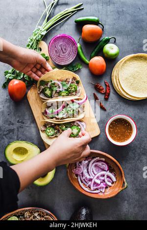 Mani latine che preparano tacos messicani con carnitas di maiale, avocado, cipolla, coriandolo e salsa rossa in Messico Foto Stock