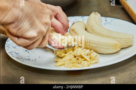Lo chef macina la banana con forchetta per fare la torta nella cucina di casa Foto Stock