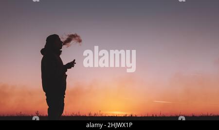 Un uomo in giacca fuma una sigaretta elettronica all'aperto. Vaporizzazione su sfondo tramonto. Alternativa alle sigarette analogiche Foto Stock