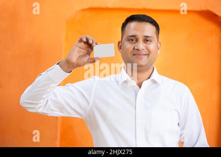 Giovane uomo indiano che mostra bianco carta mockup su sfondo arancione, sorridente ragazzo asiatico tenendo vuoto business o carta elettronica per il pagamento, de Foto Stock