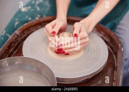 le mani delle donne con un bel lavoro di manicure rosso sulla ruota di un vasaio, scolpiscono una ciotola di argilla cruda sporca Foto Stock