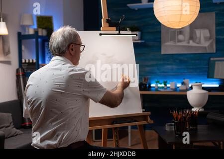 Uomo anziano disegno originale matita disegno di vaso in gesso sensazione creativa su cavalletto in casa studio. Artista in pensione che crea un autentico disegno di disegno in studio luce durante il laboratorio serale. Foto Stock