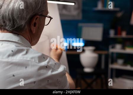 Concentratevi sul disegno di un artista in pensione su carta utilizzando la matita guardando il modello di vaso bianco in gesso in laboratorio creativo. Uomo anziano che disegna ancora oggetto facendo studio leggero in studio d'arte. Foto Stock