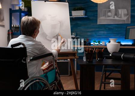 Insegnante di arte pensionato focalizzato usando la sedia a rotelle che fa l'arte creativa in matita disegno vaso modello su cavalletto. Donna anziana che vive con disabilità sensazione ispirato ombreggiatura schizzo in classe serale. Foto Stock