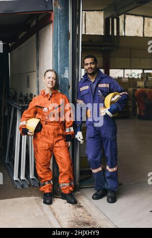 Sorridenti colleghi maschi e femmine in abbigliamento da lavoro protettivo presso il magazzino di fabbrica Foto Stock