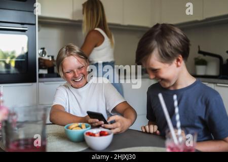 Sorridendo i fratelli usando lo smart phone mentre la madre fa i chores della cucina in background nel paese Foto Stock