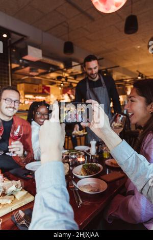 Immagine ritagliata di un uomo che fa foto di amici maschi e femmine attraverso il telefono cellulare in bar Foto Stock