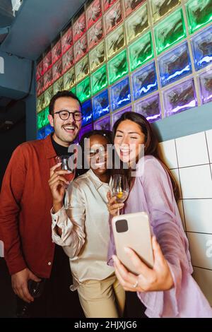 Donna allegra che prende selfie con amici maschi e femmine attraverso lo smartphone al bar Foto Stock