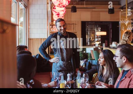 Cameriere sorridente che parla con amici maschi e femmine al ristorante durante la cena Foto Stock