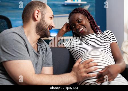 Felice gravidanza multietnica giovane coppia sorridente l'un l'altro mentre futuro padre toccare donna ventre. Allegro uomo caucasico sensazione di nascere in movimento all'interno di gioiosa donna incinta afroamericana Foto Stock