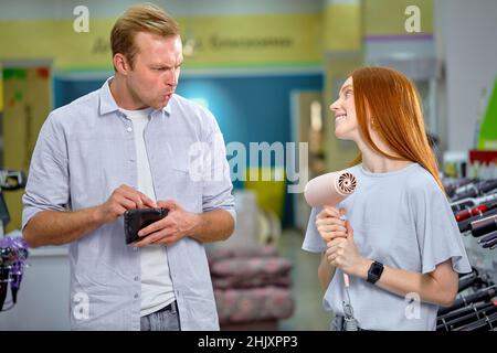 Cute Redhead femmina chiedere a marito da asciugacapelli, elemosina. L'uomo non ha soldi, tenendo la borsetta nelle mani, al negozio di elettrodomestici. Adorabile Woma adulto Foto Stock