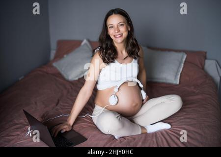 Donna incinta felice rilassarsi a casa. È seduta sul letto in camera da letto e suona la musica al suo bambino nello stomaco. Foto Stock