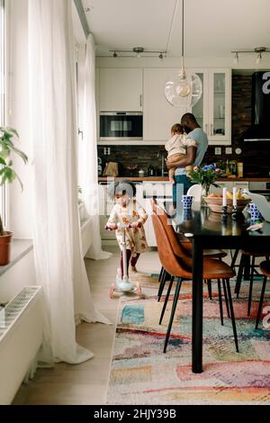 Il padre che porta il bambino ragazzo mentre la figlia che guida lo scooter a spinta in cucina Foto Stock