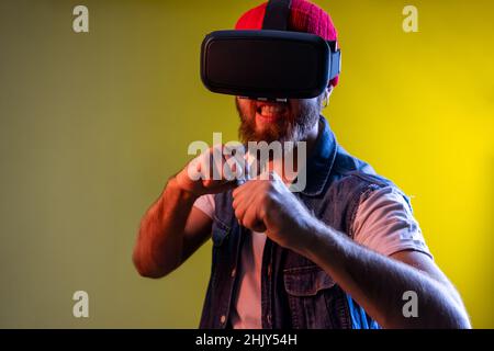 Hipster Guy in virtual reality headset con pugni stretti pronti per pugilato, gioco online, denti stretti con rabbia. Studio interno girato isolato su colorato sfondo di luce al neon. Foto Stock