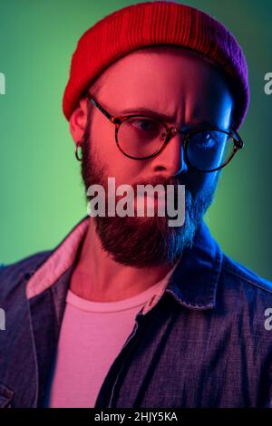 Ritratto closeup di un uomo hipster pensivo in occhiali, in piedi guardando lontano, esprimendo la pensilosità, indossare berretto di beanie e gilet denim. Studio interno girato isolato su colorato sfondo di luce al neon. Foto Stock