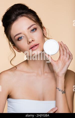 Giovane donna in contenitore bianco con crema cosmetica isolata su beige Foto Stock