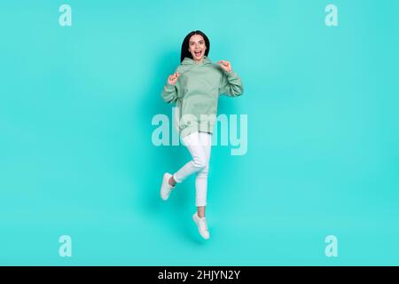 Foto in formato integrale di una bruna millenaria impressionata donna salto indossare felpa con cappuccio pantaloni scarpe isolate su sfondo teal Foto Stock