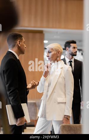 uomo di sicurezza afro-americano con computer portatile che parla con la signora anziana premurosa vicino bodyguard birazziale in hotel Foto Stock
