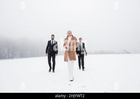 vista a tutta lunghezza dell'elegante uomo d'affari anziano che cammina attraverso il campo invernale con le guardie del corpo interrazziali Foto Stock