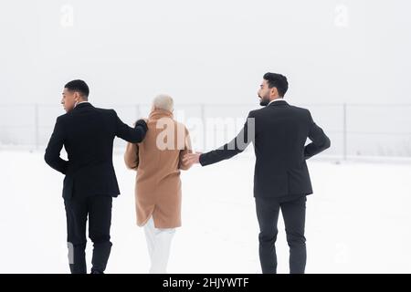 vista posteriore della donna d'affari matura che cammina attraverso il campo nevoso sotto la protezione di bodyguard interrazziali in tute nere Foto Stock