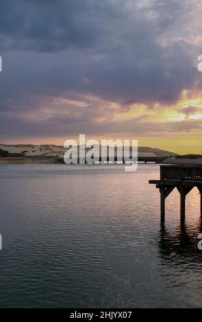 Tramonto su un lago salato. Nel lago avanza un pontile in legno, in lontananza un ponte attraversa l'acqua e va verso le dune. Foto Stock