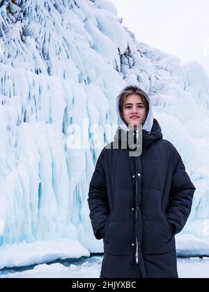 Adolescente sullo sfondo di una parete di ghiaccio, sul lago d'inverno Baikal, indossando abiti casual con un cappuccio. Verticale. Foto Stock