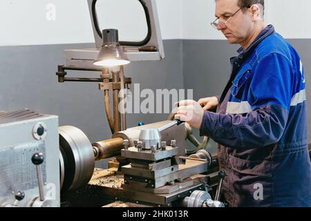 Il torcente adulto di 50-55 anni lavora in officina dietro il tornio. Produzione industriale di parti metalliche. Lavoratore caucasico in tute. Autentico processo di lavoro scena in produzione. Lavoratore reale. Scena non in scena. Foto Stock