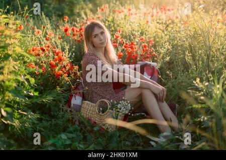 Donna bionda all'aperto in plaid con fiori di papavero al tramonto. Donna in abito e borsa con fiori da campo Foto Stock